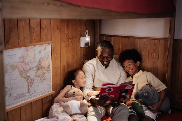 Warm, intimate scene of an adult reading a bedtime story to two children inside a wooden bunk bed. The children hold stuffed animals, and a soft wall lamp casts a gentle glow. A world map pinned to the wall adds an educational touch to the cozy setting.