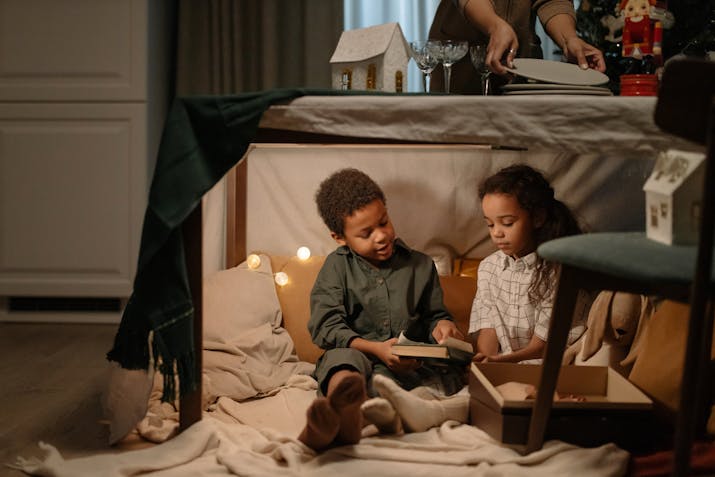 Two children sit inside a cozy indoor fort made of blankets and furniture, softly lit by string lights. One child reads a book while the other watches. In the background, an adult sets a table near a decorated Christmas tree, capturing a heartwarming holiday scene of quiet play and family togetherness.