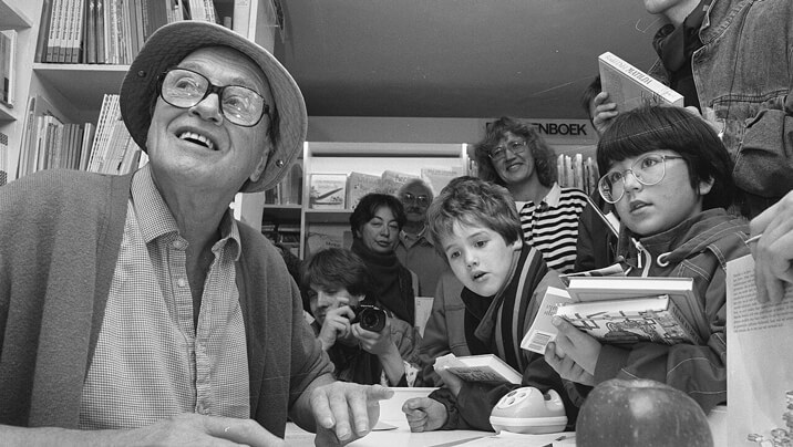 Black-and-white photo of Roald Dahl seated at a table during a book signing, surrounded by children and adults in a bookstore. He wears a hat and glasses, smiling warmly with books and an apple in front of him. Shelves filled with books and a Dutch sign reading “BOEK” appear in the background.