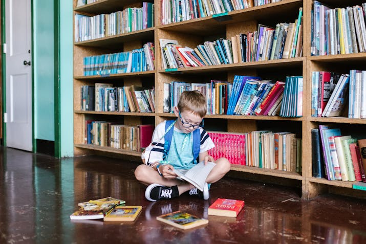 Focused child wearing glasses sits on a polished wooden floor in front of a large bookshelf, reading a book. Several other books are scattered around, and the green-and-white walls add warmth to the library-like setting.