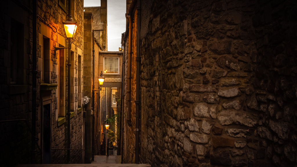 Narrow alley with stone walls and glowing street lamps at dusk.