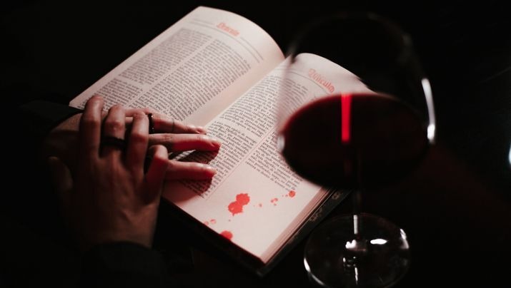 Hands reading a book with a glass of red wine nearby.